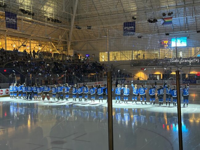 The entire PWHL Toronto team lines up at the blue line at the Mattamy Athletic Centre. The lights are low and their sticks are in the air, saluting the crowd.