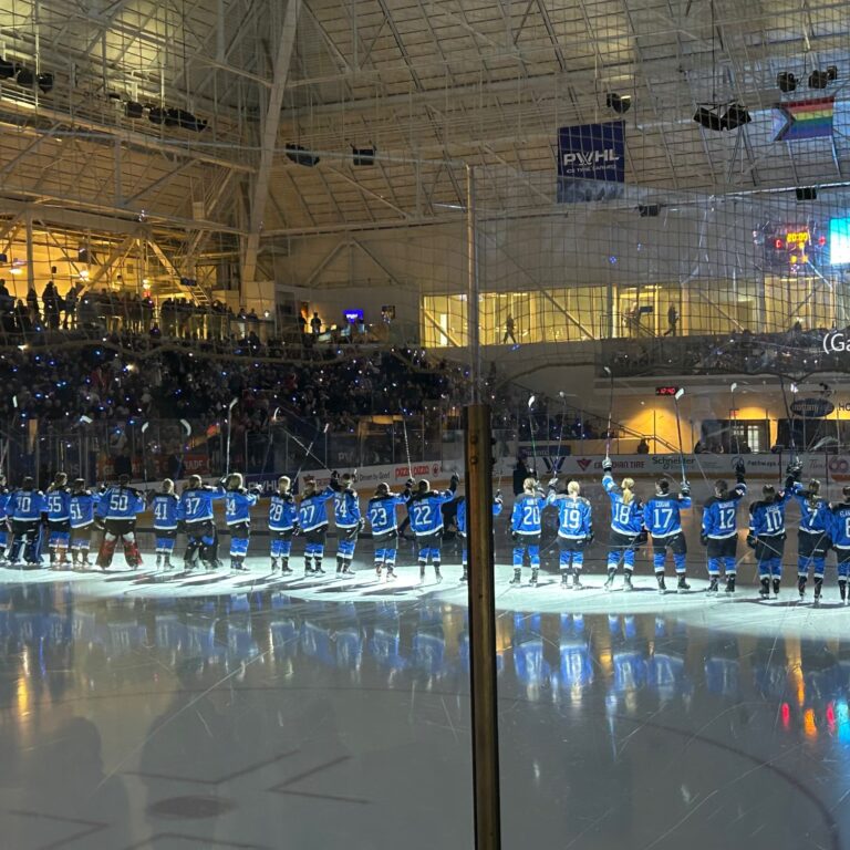 The entire PWHL Toronto team lines up at the blue line at the Mattamy Athletic Centre. The lights are low and their sticks are in the air, saluting the crowd.