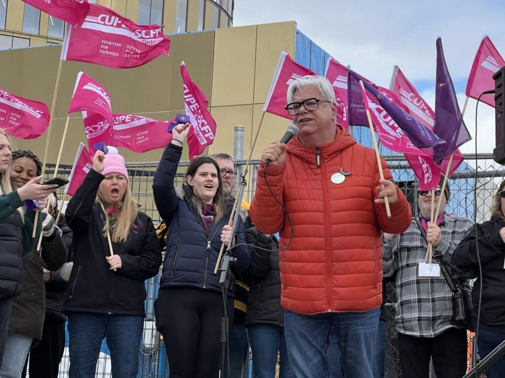 Older male speaking in-front of a crowd at a protest with individuals behind holding "CUPE 3903" flags.