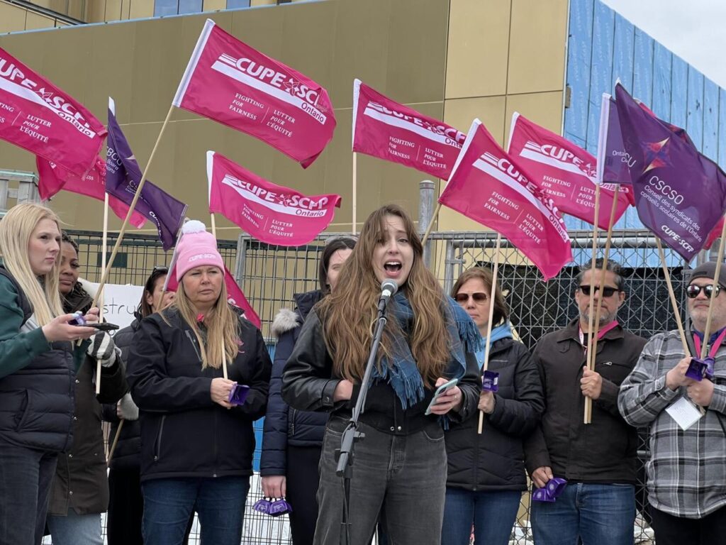 Female speaking in-front of a group of people holding "CUPE 3903" flags.