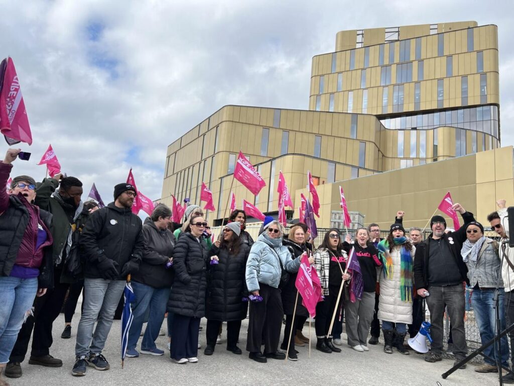 Group of people standing together holding "CUPE 3903" flags in-front of a school campus.