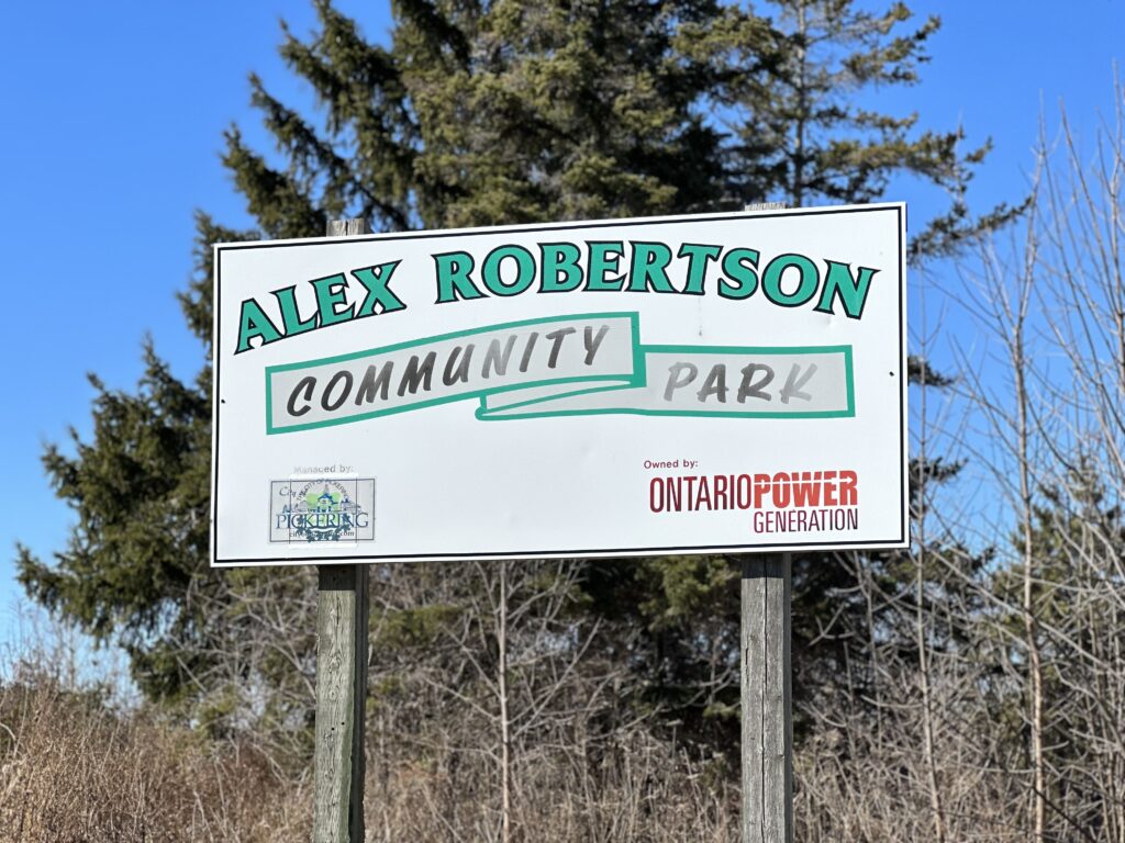 Sign reading Alex Robertson Community Park, with an Ontario Power Generation logo taking up most of the bottom right of the sign. The sign is held up by faded wooden posts, and in the background trees are visible.