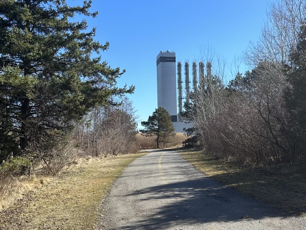 Walking path with evergreen trees along the left and bushes along the right. Further down the path, a tall portion of the nuclear station is visible.