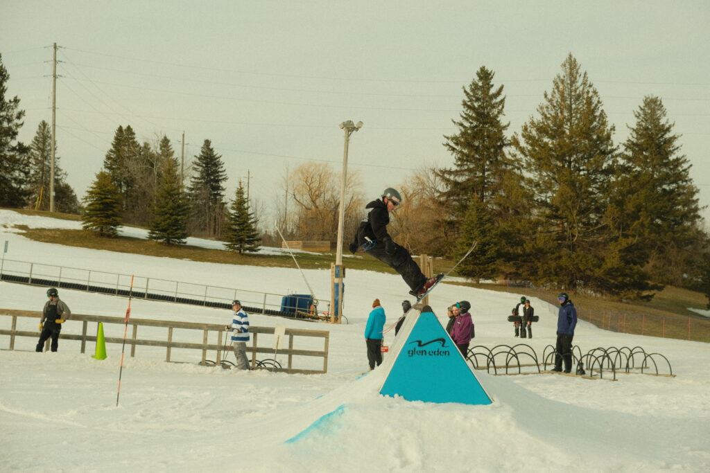 Freestyle skier rides up a triangle terrain park feature. Snow is melting in the background and patches of grass can be seen coming through the snow.