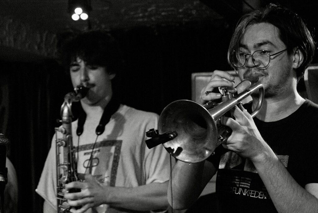 Image of a person in front playing the trumpet and someone else behind playing the saxophone.