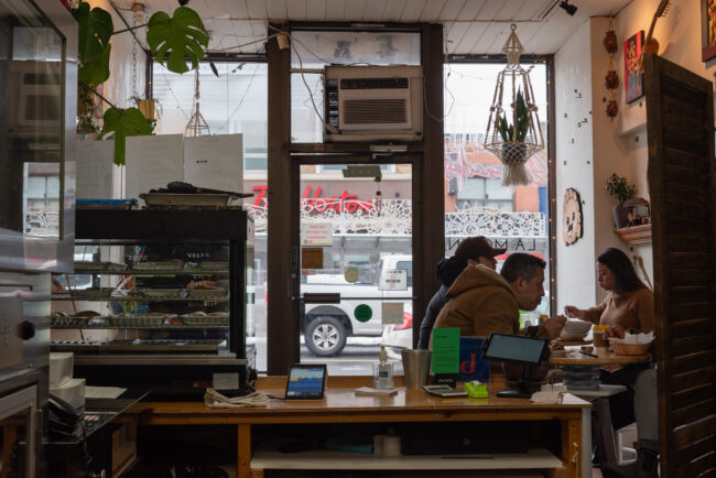 Interior of a café looking to road-facing windows. Three people sit eating at table on right.