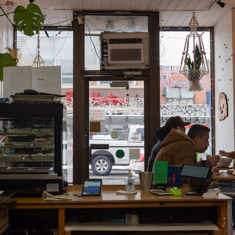Interior of a café looking to road-facing windows. Three people sit eating at table on right.