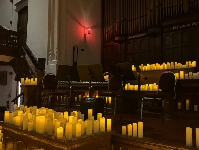 Four chairs and four music stands surrounded by large groups of LED candles placed in a dim wooden church.