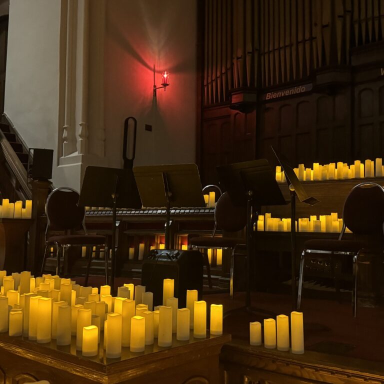 Four chairs and four music stands surrounded by large groups of LED candles placed in a dim wooden church.