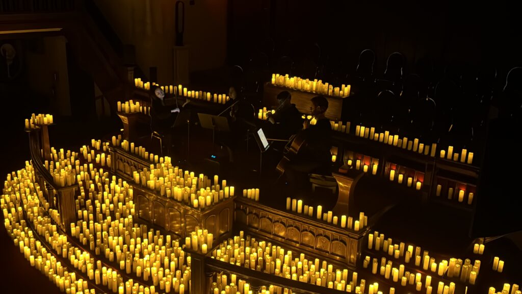 Four people playing violins and cello instruments around a large group of LED candles placed in a wooden church.