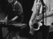 Black and white photo of a saxophone being played next to a a person playing a keyboard.
