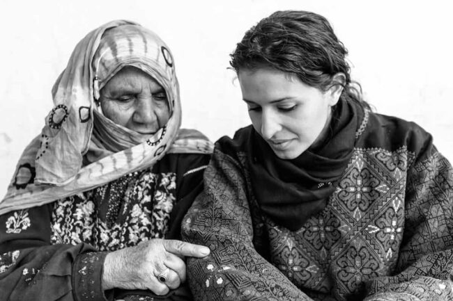 An elderly refugee woman points to the intricate embroidery on the traditional Palestinian attire worn by a younger woman, engaging in conversation.