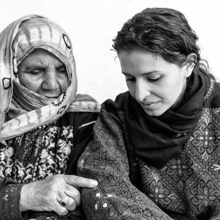 An elderly refugee woman points to the intricate embroidery on the traditional Palestinian attire worn by a younger woman, engaging in conversation.