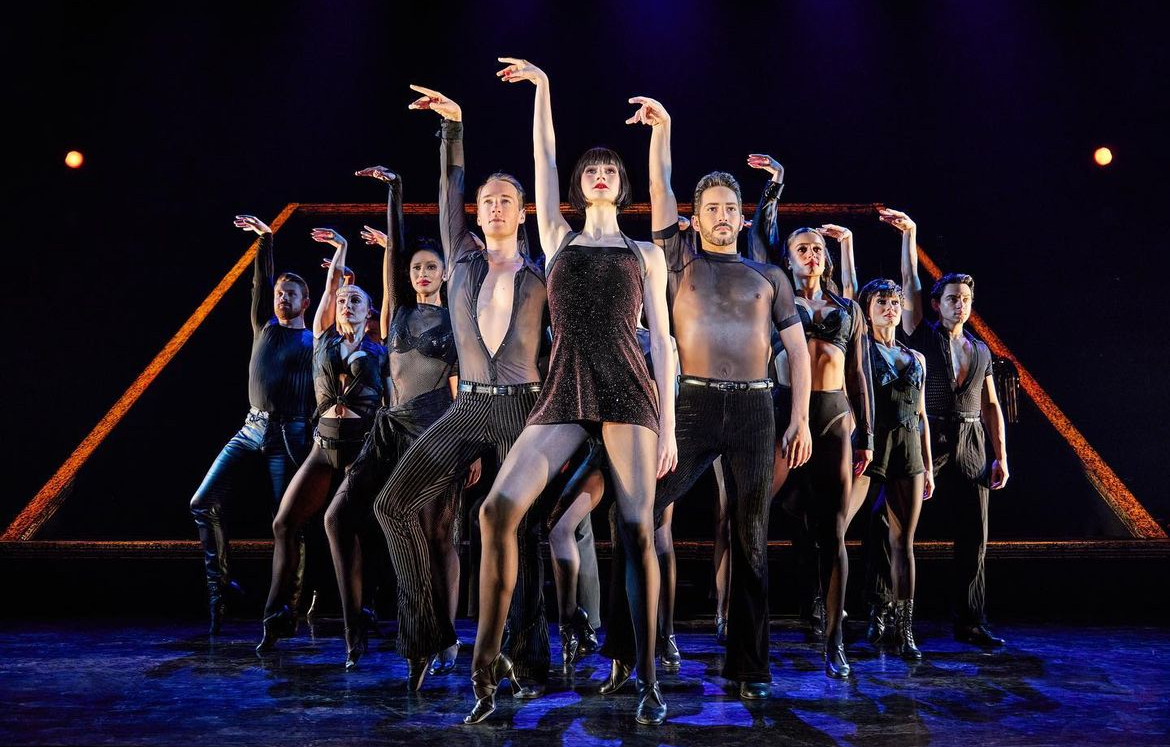 Kailin Brown is standing in the front tip of a pyramid made by dancers for a number in the musical Chicago. Each dancer has their left hand raised and their wrist is flexed. Everybody has their left leg propped up.