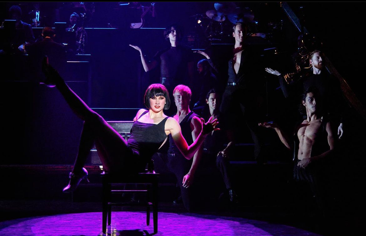A still of Kailin Brown sitting on a chair with one hand raised in the air and doing a jazz hand, and one leg kicked into the air. There are dancers in the background, some are standing, others are sitting down. They have one arm raised at a ninety degree angle with their wrist flexed.