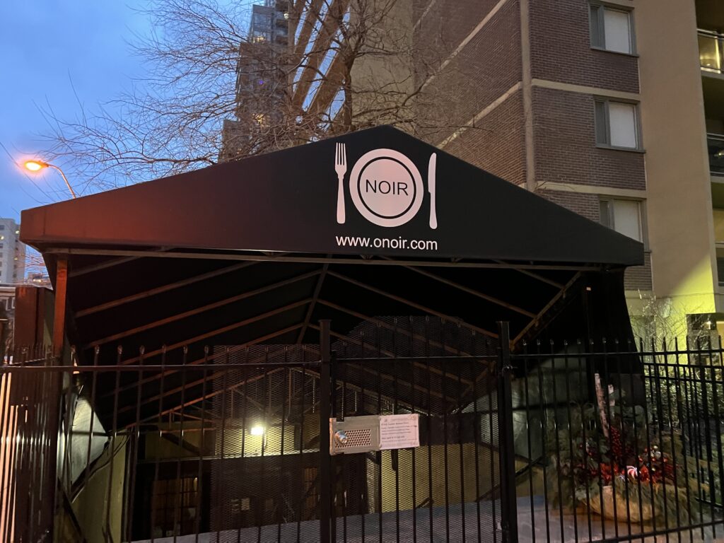 Photo of the front entrance of O'Noir Toronto. Large black tent-appearing roof with O'Noir logo of fork and knife with a plate in the centre with text "Noir" in the middle. The tent is over stairs going down to the main door, with an iron gate in front.  