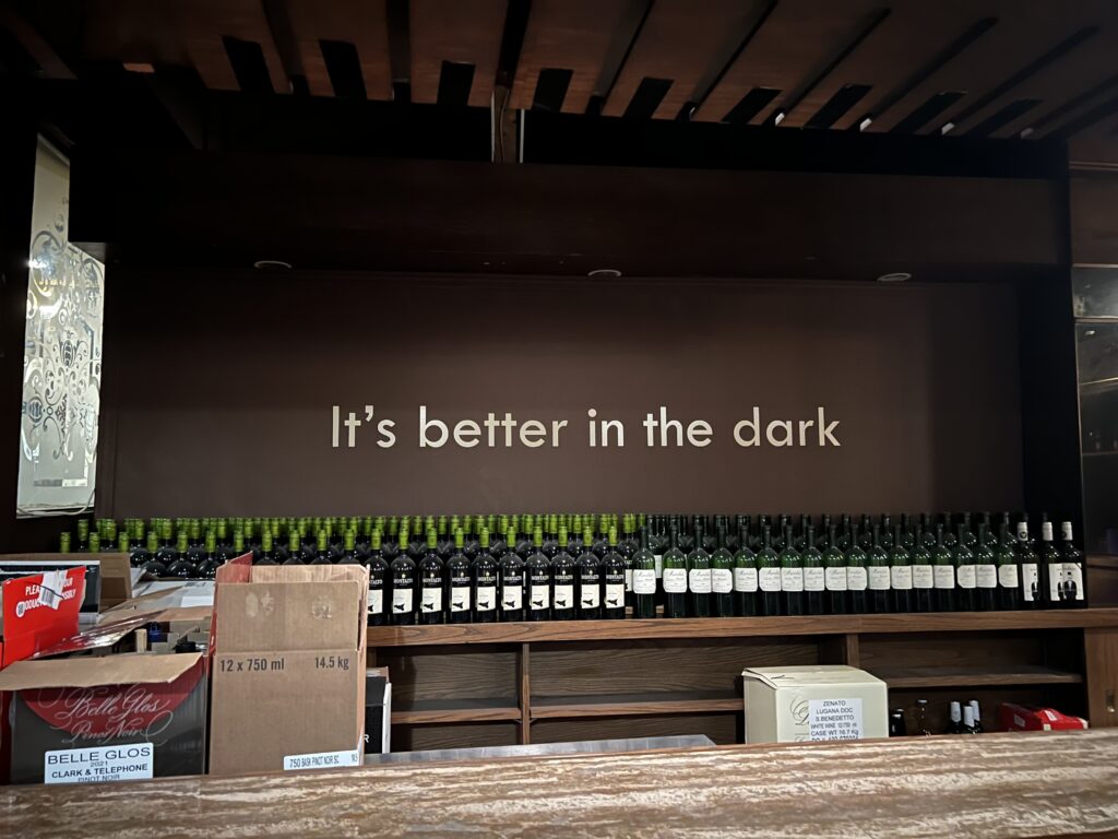 A photo of the bar area of O'noir. The wall is painted brown with "It's better in the dark" written in beige paint. The bar area is lined with many bottles of wine. 