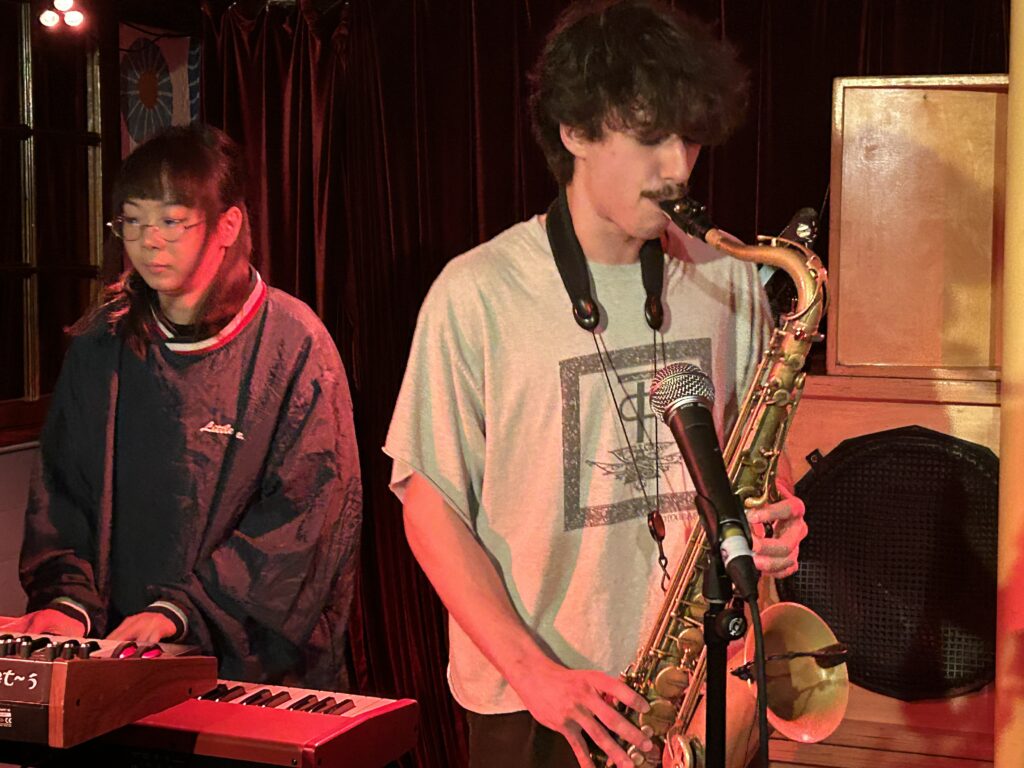 Image of someone playing the saxophone and someone behind playing the keyboard.