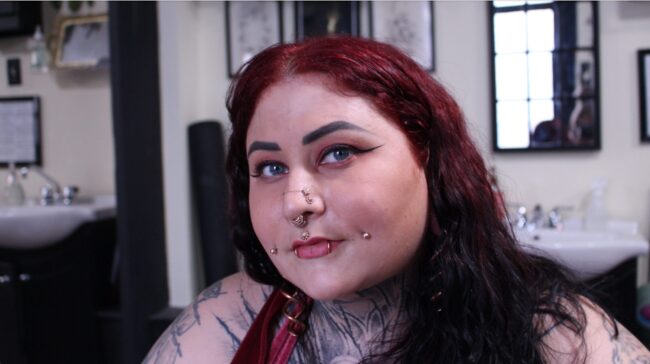 A woman with bright red hair and multiple piercings and tattoos gazes into the camera