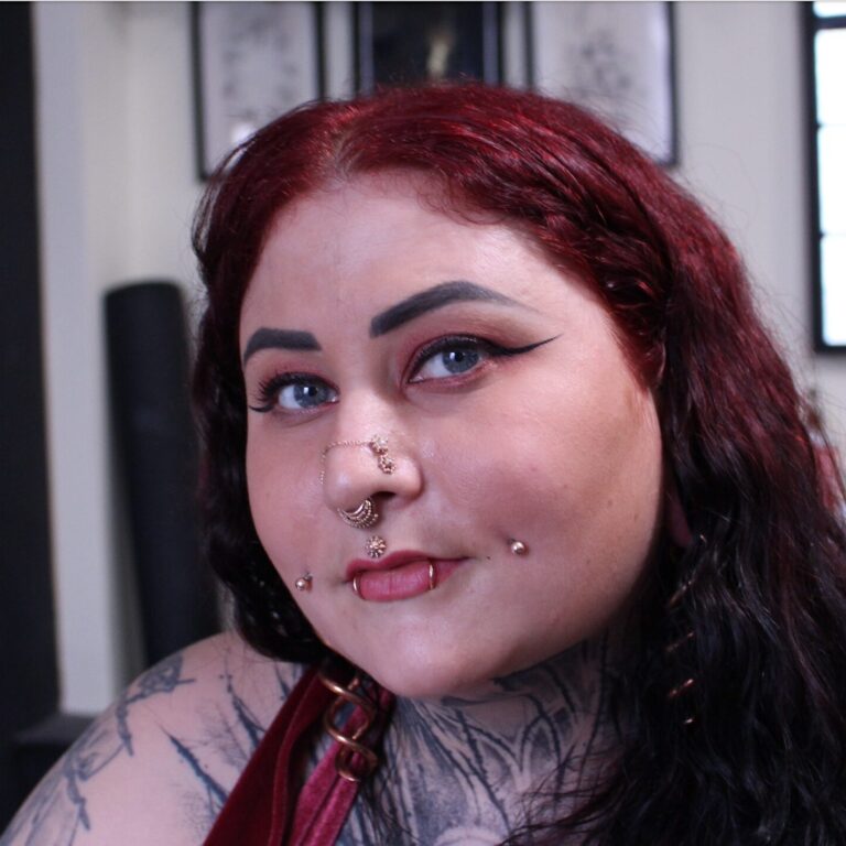 A woman with bright red hair and multiple piercings and tattoos gazes into the camera