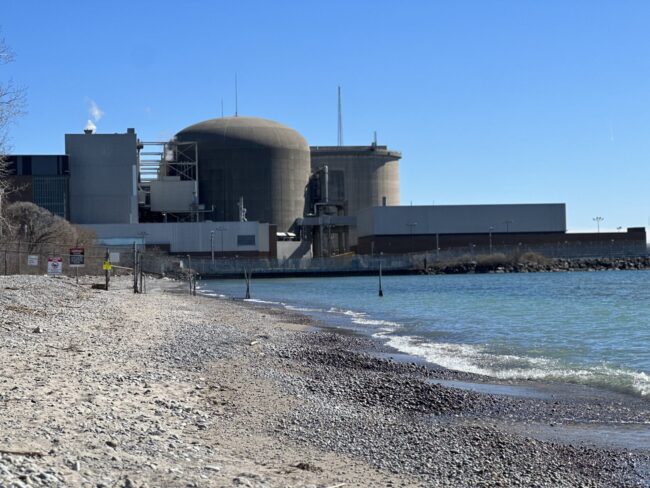 View of a beach with rocky sand on the left and waves on the right. In the background, a nuclear power plant is visible, blocked by a fence marked with caution signs.