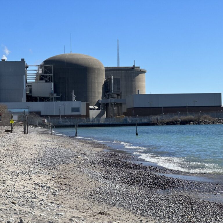 View of a beach with rocky sand on the left and waves on the right. In the background, a nuclear power plant is visible, blocked by a fence marked with caution signs.