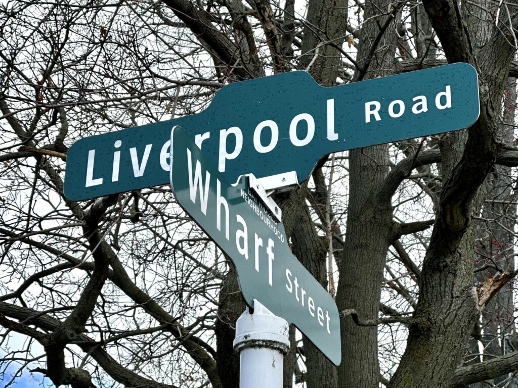 Green road signs in front of a tree. The one on top reads "Liverpool Road". The one on bottom reads "Wharf Street."