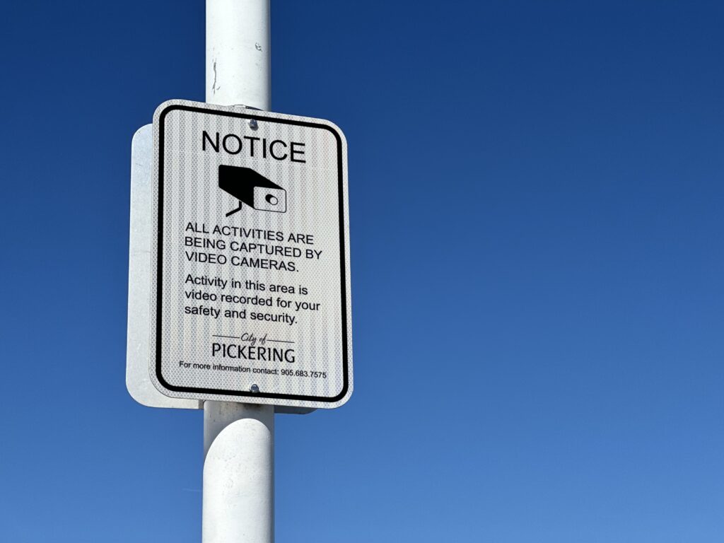 A sign against blue sky with a picture of a security camera. It reads "Notice. All activities are being captured by video camera. Activity in this area is video recorded for your safety and security. With the logo of the City of Pickering.