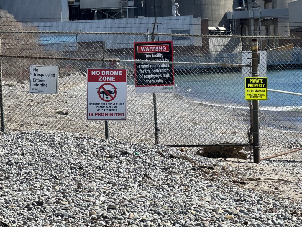 Four caution signs adorning a barbed wire-topped fence. One warns against trespassing, one against drone and video recording. One indicates that the area is monitored 24/7 by armed guards. The last one states that this is private property. The nuclear plant is just visible in the backround, and the bottom portion of the photo is a rocky beach area leading up to the fence.