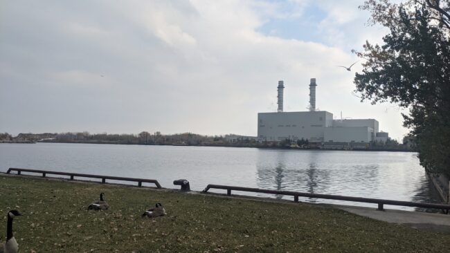 A photo of the Portlands Power Plant in downtown Toronto. The Portlands Power Plant is white and seems to be in operation as there is flue gas coming out of the stack. There are geese in the foreground