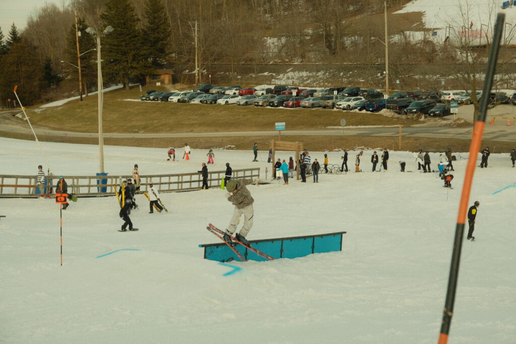 Freestyle skier rides down a rail in the terrain park. There are other freestyle skiers in the background hiking up the park. Snow is melting in the background and patches of grass can be seen coming through the snow.