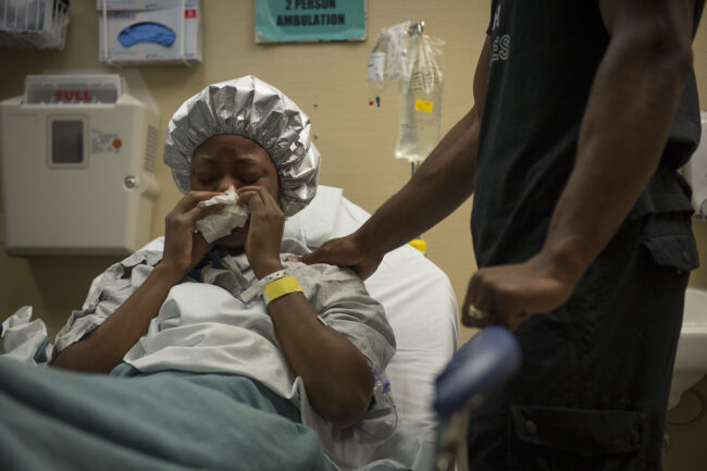 A black woman in a hospital gown being comforted while she blows her nose in bed.