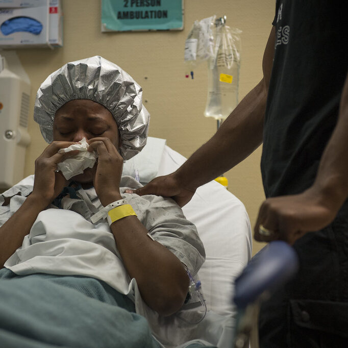 A black woman in a hospital gown being comforted while she blows her nose in bed.