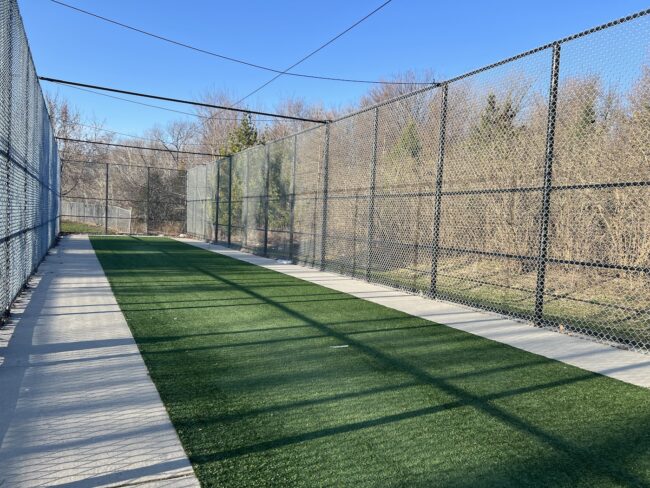 A singular turf cricket lane with a black barred fence around three sides with blue skies and trees in the background.