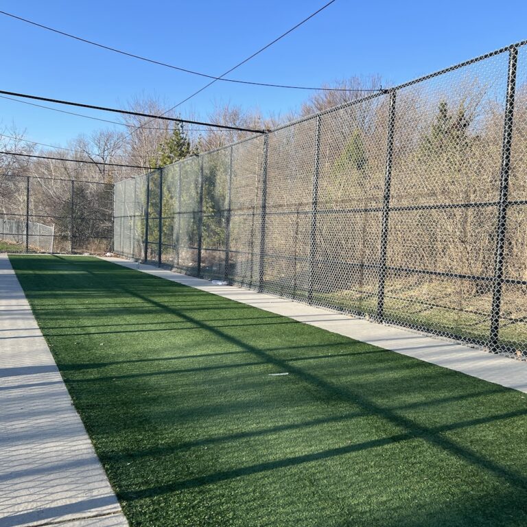 A singular turf cricket lane with a black barred fence around three sides with blue skies and trees in the background.