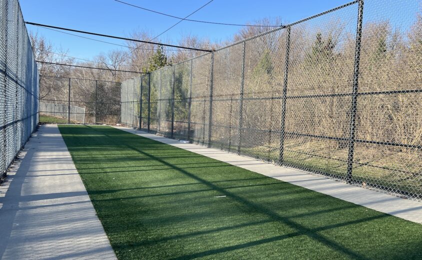 A singular turf cricket lane with a black barred fence around three sides with blue skies and trees in the background.