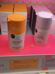 Two white skincare bottles with an orange and pink lid on a pink display shelf with brown product stains.