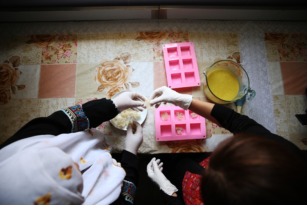 Two women carefully add an ingredient into the container, preparing for the soap mixture.