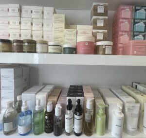 A white shelf with colourful jars of toner pads on the top shelf and bottles of cleansing oil at the bottom-level of the shelf.