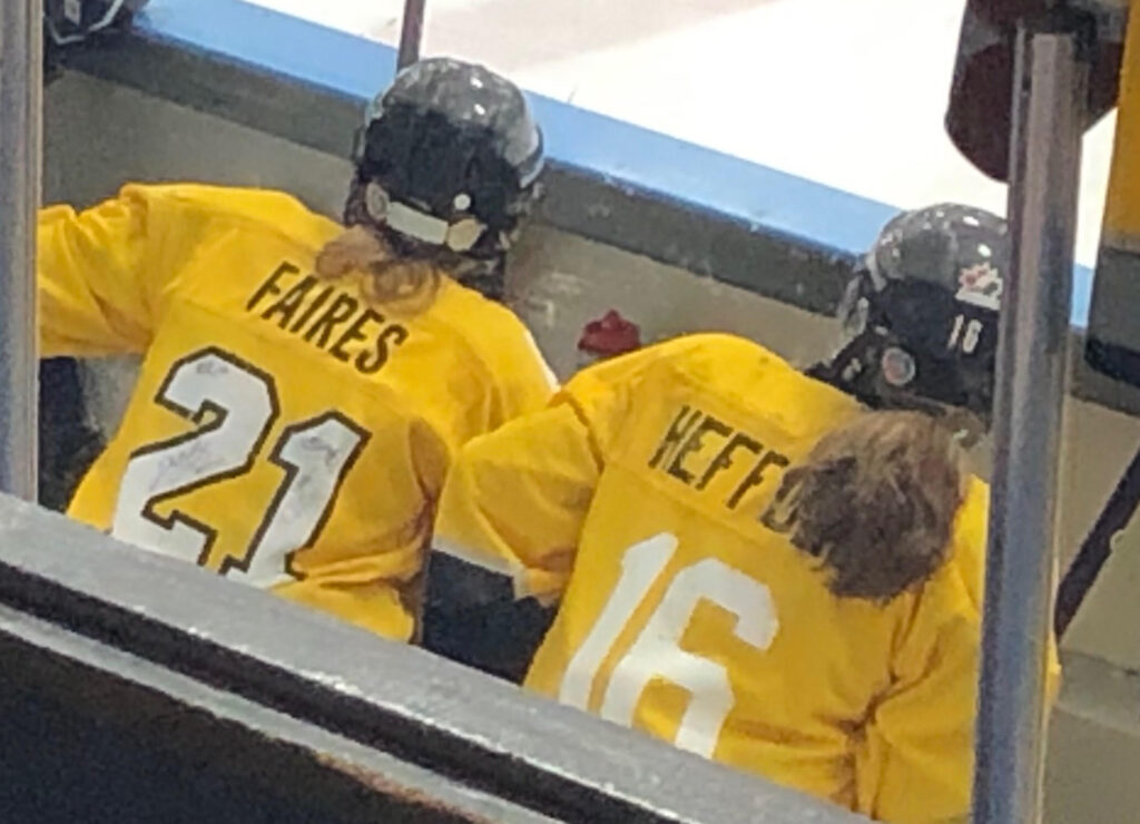 Jennifer Faires sitting beside Jayna Hefford on a hockey bench. Both are wearing yellow jerseys and black helmets. Their last names "Faires" and "Hefford" are visual on the back of their jerseys.